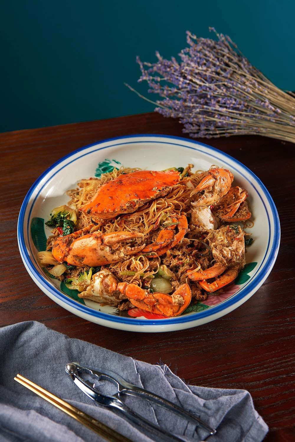 Black Pepper Crab Bee Hoon: whole crab pieces on stir-fried bee hoon in a peppery sauce, served on a blue-rimmed plate with a crab cracker beside it