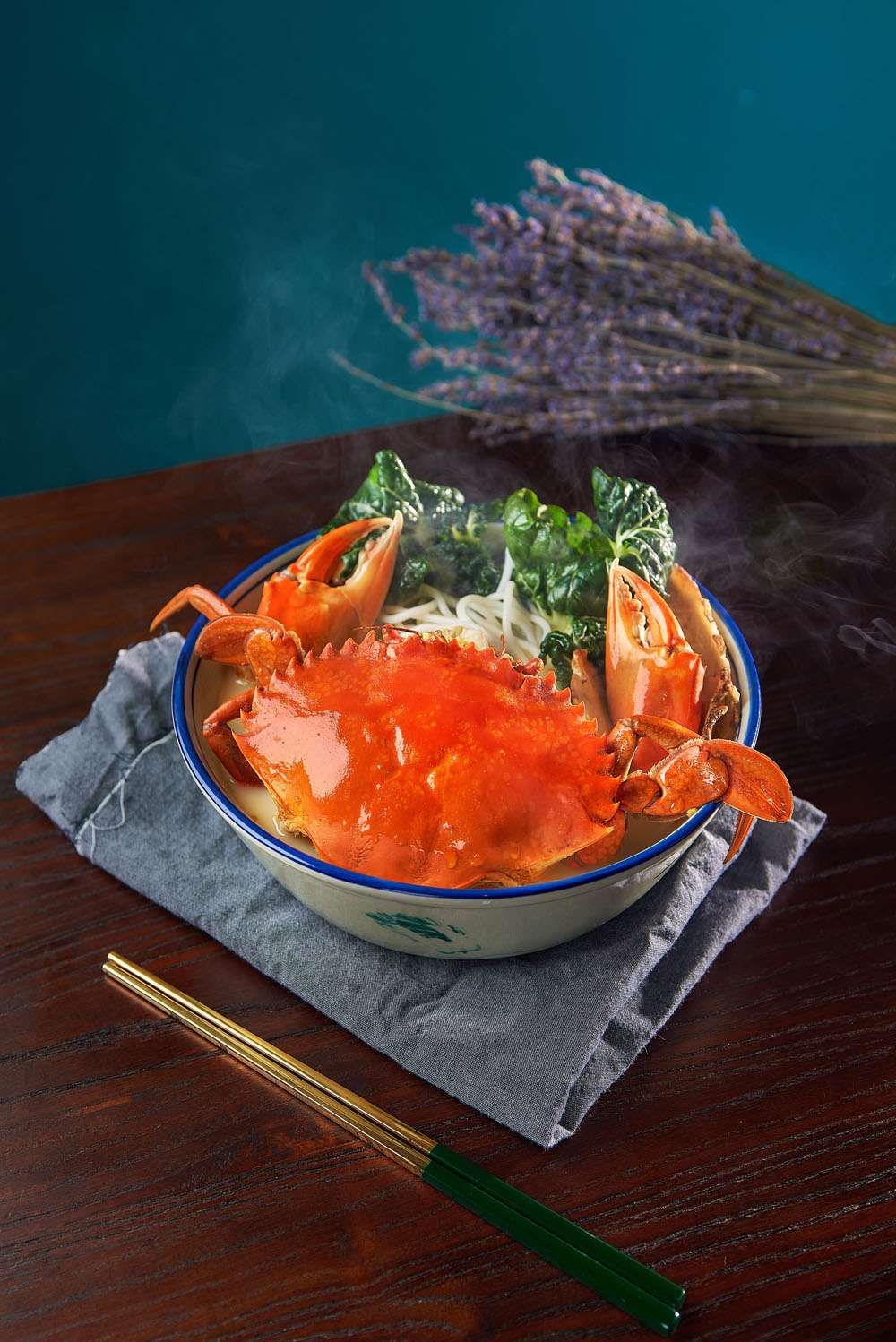 A whole Sri Lankan mud crab in milky herbal broth with bee hoon and greens in a blue-rimmed bowl, steam rising, with gold chopsticks beside it